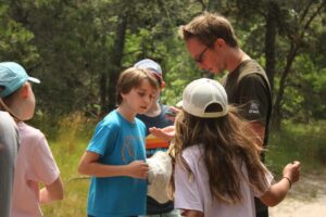 Group of children gatherd around a man looking at what is in his hand. 